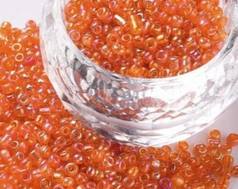 May include: A collection of small, round, translucent orange glass seed beads with an iridescent finish. The beads are contained within a clear, faceted glass bowl, with a large pile of identical loose beads surrounding the base of the bowl.