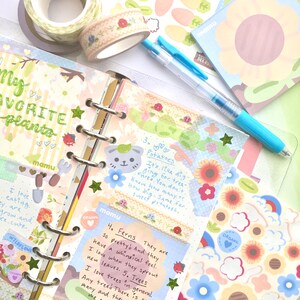 K&ouml;nnte beinhalten: Ein Planer mit floralen und botanischen Dekorationen. Er enth&auml;lt Aufkleber, Washi-Tape und handgeschriebene Notizen &uuml;ber Pflanzen. Ein hellblauer Stift liegt auf den offenen Seiten. Der Text "My Favorite Plants" ist sichtbar.