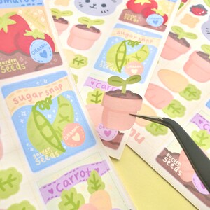 K&ouml;nnte beinhalten: Eine Sammlung farbenfroher Stickerb&ouml;gen mit Illustrationen zum Thema Garten. Designs umfassen Saatgutt&uuml;ten mit Texten wie "sugarsnap" und "carrot" sowie Abbildungen von Tomaten, Erbsen und Karotten. Die Sticker haben ein "organic" Herz-Logo.
