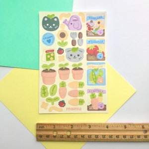 K&ouml;nnte beinhalten: Ein Bogen bunter Aufkleber mit einem Gartenthema. Die Aufkleber zeigen Illustrationen einer Gie&szlig;kanne, Topfpflanzen, Gem&uuml;se und niedlichen Tiergesichtern. Die Texte "ORGANIC" und "mamu" sind sichtbar. Das Blatt ist etwa 10 cm breit.