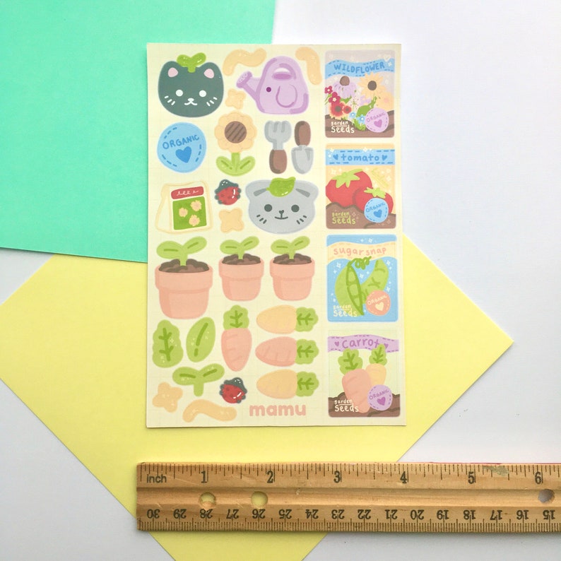 K&ouml;nnte beinhalten: Ein Bogen bunter Aufkleber mit einem Gartenthema. Die Aufkleber zeigen Illustrationen einer Gie&szlig;kanne, Topfpflanzen, Gem&uuml;se und niedlichen Tiergesichtern. Die Texte "ORGANIC" und "mamu" sind sichtbar. Das Blatt ist etwa 10 cm breit.