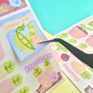 K&ouml;nnte beinhalten: Ein Bogen mit farbenfrohen Aufklebern zum Thema Garten. Ein Aufkleber zeigt ein Zuckerschoten-Design mit dem Text "sugar snap" und "garden seeds". Ein weiterer Aufkleber zeigt Karotten mit dem Text "carrot" und "garden seeds". Die Aufkleber haben ein herzf&ouml;rmiges "organic"-Etikett.