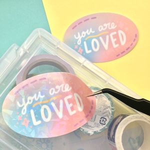 May include: Two oval stickers with a holographic finish and the text "you are loved" in pink, blue, and yellow lettering. The stickers are in a clear plastic container with other craft supplies.