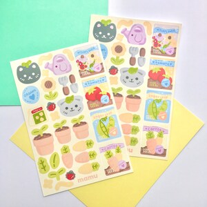 K&ouml;nnte beinhalten: Zwei Stickerb&ouml;gen mit Illustrationen zum Thema Garten. Designs umfassen ein Katzen-Gesicht, eine Gie&szlig;kanne, eine Sonnenblume, Tomaten, Karotten und Sament&uuml;ten. Die Aufkleber tragen die W&ouml;rter "ORGANIC" und "mamu".