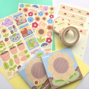 K&ouml;nnte beinhalten: Eine Sammlung von Schreibwaren mit Gartenthema. Enth&auml;lt Stickerb&ouml;gen mit Gartenillustrationen, dekorative Klebebandrollen und bl&uuml;tenf&ouml;rmige Notizbl&ouml;cke. Die Farbpalette umfasst Pastellt&ouml;ne von Gr&uuml;n, Gelb und Blau.