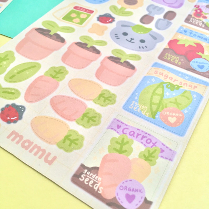 K&ouml;nnte beinhalten: Ein Bogen mit bunten Aufklebern mit Illustrationen zum Thema Garten. Designs umfassen Karotten, Topfpflanzen, Saatgutt&uuml;ten mit der Aufschrift "tomato", "sugarsnap" und "carrot" sowie ein niedliches Katzen-Gesicht. Das Wort "mamu" ist unten links aufgedruckt.