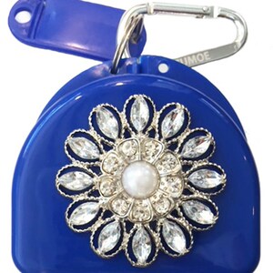 May include: A blue plastic dental retainer case with a silver and rhinestone flower design. The case has a blue clip with the word "UMOE" on it.