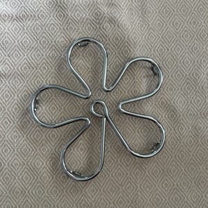 May include: A silver metal trivet shaped like a five-petal flower. The trivet is constructed from curved metal rods and is designed to protect surfaces from hot cookware. The background is a beige fabric with a diamond pattern.