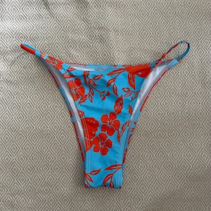 May include: A blue bikini bottom with an orange floral pattern. The bikini bottom has a high-cut design with thin straps and a white lining. The floral pattern features large flowers and leaves.