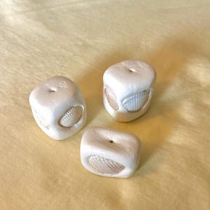 May include: Three off-white, cube-shaped beads with rounded edges, each featuring a small, centred hole on the top surface. Each bead has a light tan, textured seashell embedded into one of its sides, creating a contrast between the smooth matte finish of the bead and the natural ridges of the shell.