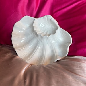 May include: A white ceramic serving dish with a petal design. The dish features a large, shallow bowl with a smaller, raised section in the centre. The surface is smooth and glossy, reflecting light. It is likely used for serving food or as a decorative piece.
