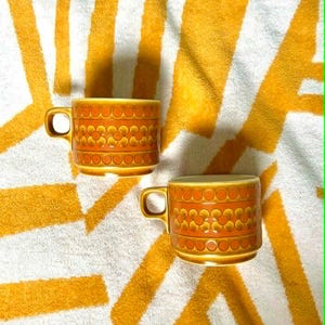 Puede incluir: Dos tazas de cerámica naranja con un diseño floral y asas pequeñas. Las tazas están sobre un fondo a rayas blancas y amarillas, creando una estética retro. Las tazas son probablemente para café o té.