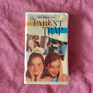 Puede incluir: Una cinta VHS de la película The Parent Trap, con el título en texto negro, un logotipo de Walt Disney Pictures y tres escenas fotográficas de la película, incluyendo dos niñas idénticas de cabello rojizo apoyando sus rostros en sus manos.