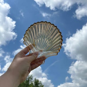May include: A clear, seashell-shaped dish with a scalloped edge. The dish has a ribbed design and a golden-brown rim. The background is a bright blue sky with fluffy white clouds.