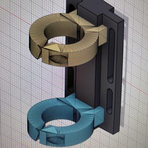 Op de afbeelding: Een 3D-geprint zwart plastic beugel met twee ringen. De bovenste ring is beige en de onderste ring is blauw.