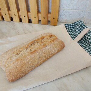 Peut inclure: Un pain rectangulaire à la croûte dorée est posé sur un sac en tissu beige. Le sac présente une fermeture à cordon avec un motif à carreaux verts et blancs visible à l'ouverture, ainsi qu'une boucle en tissu assortie fixée sur le côté.