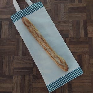 Peut inclure: Un sac en tissu rectangulaire de couleur blanc cassé, conçu pour transporter une baguette. Le sac présente un motif à carreaux verts et blancs sur les bords supérieur et inférieur, avec une anse en tissu. Une baguette dorée est posée dessus.