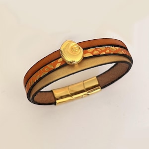 Peut inclure: Bracelet en cuir à plusieurs brins avec un fermoir coquillage doré. Le bracelet présente des bandes de cuir marron, dorées et orange. La bande orange a un motif de feuilles dorées, et le fermoir est magnétique.