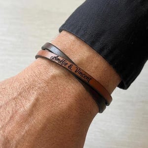 May include: A double-wrap leather bracelet featuring a light brown strap and a dark brown strap. The light brown strap is engraved with the black text Amélie & Vincent in a cursive script font. The bracelet is worn around a wrist.