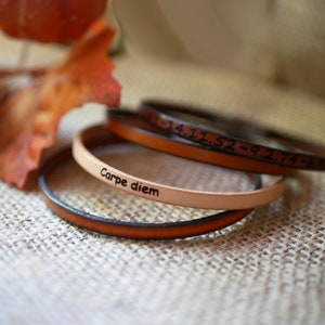 May include: Three leather bracelets stacked on top of each other. The top bracelet is brown and has the numbers "3.14.1.52 - 9.274" engraved on it. The middle bracelet is tan and has the words "Carpe diem" engraved on it. The bottom bracelet is brown.