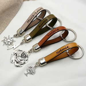 May include: Four leather keyrings with silver key rings and charms. The keyrings are in different colours: beige, green, brown, and yellow. Each keyring has a name engraved on it: Marie-Laure, Aurore, Valérie, and Noémie. The charms are a sun with a lightning bolt, a rose, and a tree.