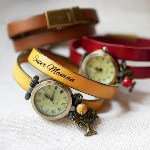 Può includere: Una collezione di orologi da polso in pelle in vari colori. Un orologio presenta un cinturino giallo senape con la scritta "Super Maman". Altri cinturini sono marroni e rossi. Ogni orologio ha un quadrante rotondo con numeri romani e ciondoli decorativi.