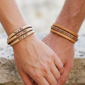 Può includere: Braccialetti in pelle color cuoio con testo inciso. Un polso mostra "Tom et Noa", "Jules et Ambre" e "Jade et Léa". L'altro braccialetto recita "comme notre espoir est en toi I (ptoune".