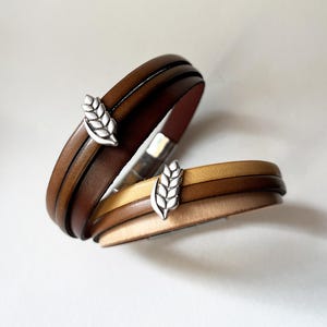 Può includere: Due braccialetti in pelle marrone e beige, ciascuno con un ciondolo a forma di spiga di grano argentata. I braccialetti hanno un design a strati e una chiusura magnetica. Un accessorio elegante.