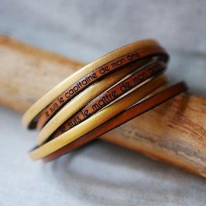 Può includere: Un set di quattro braccialetti in pelle in tonalità dorate e marroni. I braccialetti sono incisi con la frase francese "Je suis le capitaine de mon âme", che si traduce in "Sono il capitano della mia anima".