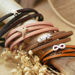 Peut inclure: Un ensemble de quatre bracelets en cuir avec différents charmes et un texte gravé. Les bracelets sont marron, beige et rose. Les charmes comprennent une étoile, un colibri, une plume et un symbole d'infini. Le texte sur les bracelets est "You are my world", "Carpe Diem", et "Infinity".