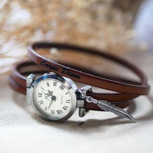 Può includere: Orologio al quarzo color argento con quadrante bianco e numeri romani, attaccato a un braccialetto avvolgente in pelle marrone. Un ciondolo a forma di piuma d'argento pende dall'orologio. Le parole "Carpe Diem" sono incise sulla pelle.