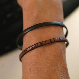 Peut inclure: Deux bracelets sur un poignet. Le bracelet supérieur est noir et lisse. Le bracelet inférieur est en cuir marron avec des coordonnées gravées : N 44° 16' 34.479 E 5° 16' 25.944.