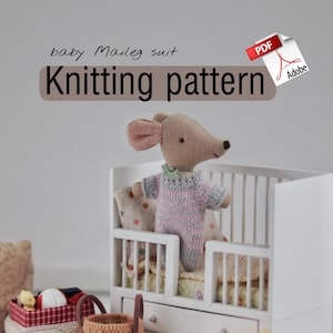May include: A miniature white crib with a knitted baby mouse wearing a pink and gray outfit. The image also features a small woven basket, a decorative box, and the text "Knitting pattern".