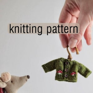 Puede incluir: Un pequeño cárdigan verde tejido con aplicaciones de setas es sostenido por una mano. Un muñeco de ratón está sentado en una mecedora roja, con una pequeña mesa y un pastel cerca. Se muestra el texto "knitting pattern".