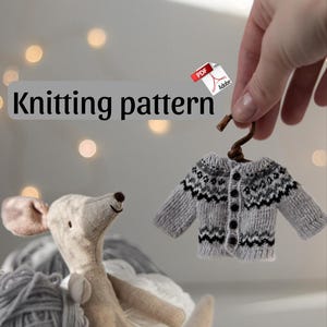 Könnte beinhalten: Ein Miniatur-Strickpullover in Grau und Weiß mit schwarzem Muster und Knopfverschluss hängt an einem kleinen Holzbügel. Ein Stoffmaus-Spielzeug sitzt neben grauen Wollknäueln. Die Worte "Knitting pattern" werden in einem Banner angezeigt.