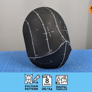 May include: A black foam helmet base featuring white hand-drawn guide lines, numerical markings, and crosshair alignment points. The object is a rounded, smooth-textured piece with a curved front edge, designed as a template for crafting.