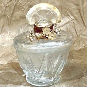 May include: A clear glass perfume bottle with a wide, faceted base and a semi-circular stopper. The neck is adorned with a dark brown velvet ribbon, metallic gold-toned floral embellishments, and small spherical beads arranged in a cluster.