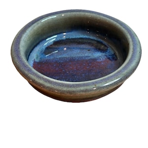 May include: A small, round ceramic dish with a glossy finish. The interior features a blend of blue and burgundy hues, while the rim is a muted gray-green. The dish has a simple, functional design, suitable for serving small portions.