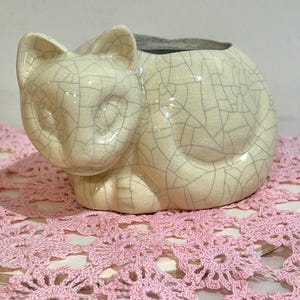 May include: A cream-colored ceramic cat-shaped planter with a crackle glaze. The cat is curled up, with its head and ears visible. The planter sits on a pink crocheted doily. A decorative item for cat lovers.