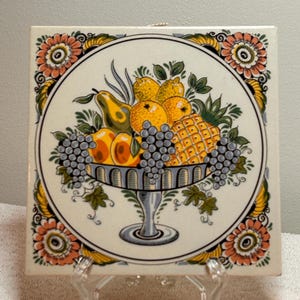 May include: Square ceramic tile featuring a detailed illustration of a fruit bowl. The bowl is filled with grapes, oranges, pears, and a pineapple. The tile is bordered by floral designs in orange, yellow, and green, set against a white background.