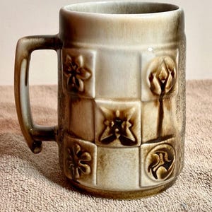 May include: A beige ceramic mug with a handle. The mug features a textured surface and is decorated with embossed floral and abstract designs within a grid pattern. The handle and rim have a darker brown color.