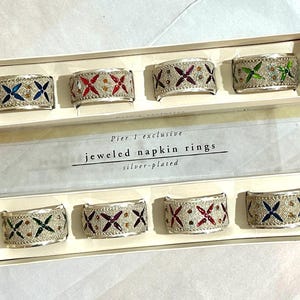 May include: Set of silver-plated jeweled napkin rings. Each ring features a decorative design with colorful jewels in blue, red, green, and brown. The rings are presented in a white box with the text "Pier 1 exclusive jeweled napkin rings silver-plated."
