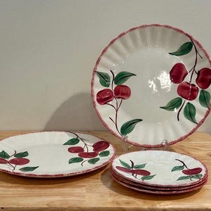 May include: A set of ceramic dinnerware featuring a cream-colored base with a scalloped edge and a red speckled rim. The pieces are decorated with hand-painted red cherries and green leaves on stems. The set includes one large plate and four smaller plates.