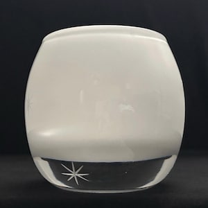 May include: A rounded glass votive candle holder with a smooth, opaque white finish on the upper portion and a clear glass base. The clear base features a delicate, etched starburst design, adding a subtle detail to the simple, curved form.