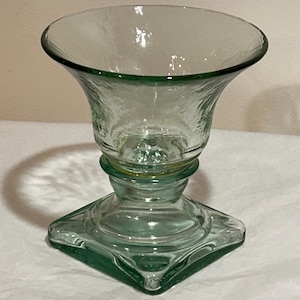 May include: A clear, green-tinted glass candle holder with a flared top and a square base. The candle holder has a textured surface and a decorative design. The overall height is approximately 6 inches.