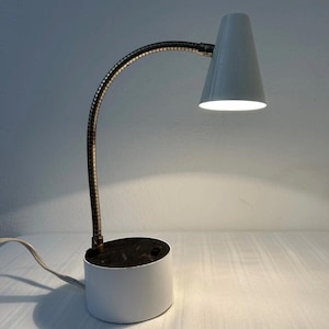 May include: A white desk lamp with a conical shade and a flexible, metallic gooseneck arm. The lamp features a cylindrical white base with a dark top surface and a power cord extending from the side. The light is illuminated, casting a glow.