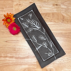 May include: A rectangular black ceramic serving tray with a white leaf design. The tray is decorated with a white leaf pattern. Two artificial flowers, one orange and one pink, are placed near the tray.