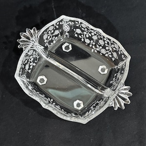 May include: A clear glass divided serving dish featuring a central divider and two handles shaped like stylized fans. The rim and sides are decorated with an etched floral vine pattern, and the base includes four small, raised hexagonal feet.