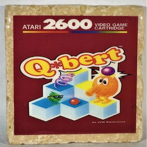 May include: A red Atari 2600 video game cartridge with the title "Q*bert" in yellow letters. The game features a cartoon character with an orange body, purple eyes, and a long orange nose. The character is standing on a blue and yellow platform with a green character with sunglasses.