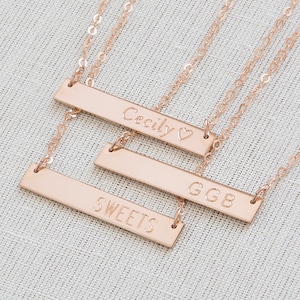 May include: Three gold bar necklaces with personalized engravings. The top necklace reads "Cecily" with a heart symbol. The middle necklace reads "GGB" and the bottom necklace reads "SWEETS".